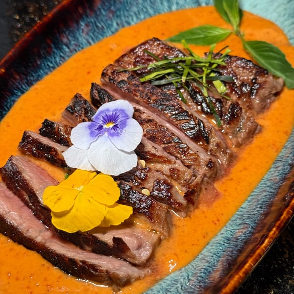 Sliced grilled beef with orange sauce, garnished with edible flowers and herbs on a blue plate.