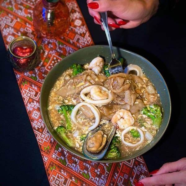 A person with red nails eating a bowl of seafood noodles with broccoli and a mussel.