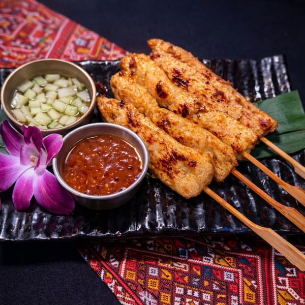 Grilled chicken satay skewers with peanut sauce, cucumber relish, and a purple orchid on a black plate.